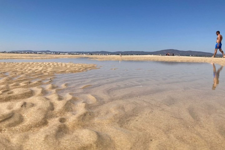 Man walking on a sandy beach with shallow water under a clear blue sky.
